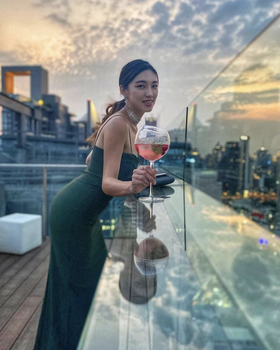 a Thai girl standing by a window panel on a rooftop in Bangkok holding a large glass of wine