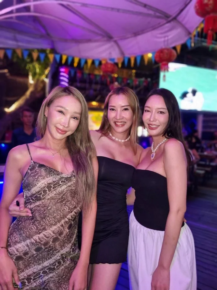 Three women in evening outfits posing together at Green Mango Club in Koh Samui with colorful bunting, red lanterns, and a DJ screen in the background.