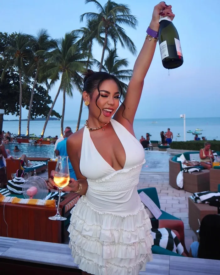A woman in a white dress raising a champagne bottle with one hand and holding a glass with the other at SEEN Beach Club in Chaweng with palm trees, the pool, and the sea at dusk in the background.