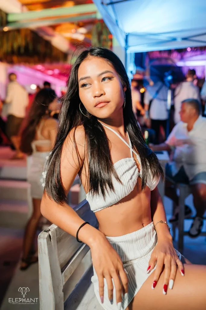A cute Thai babe in a white crop top and shorts leaning on a chair at Elephant Beach Club in Koh Samui with a lively crowd and colorful lights in the background.